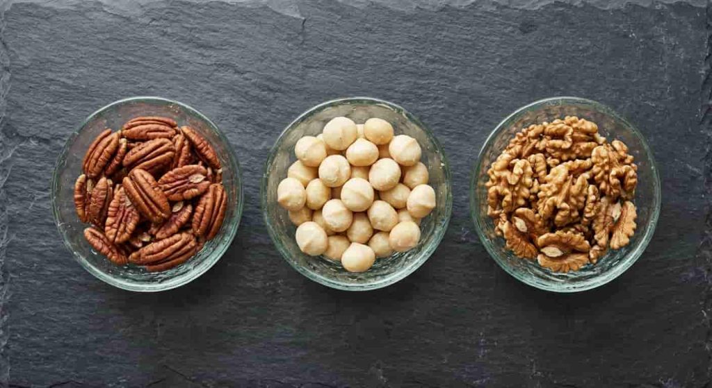 Three glass bowls filled with pecans, macadamia nuts, and walnuts representing the lowest carb nuts for keto diet.